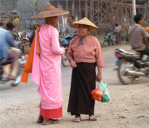 A traditionally dressed female monk and old woman stand in the street as traffic passes by.